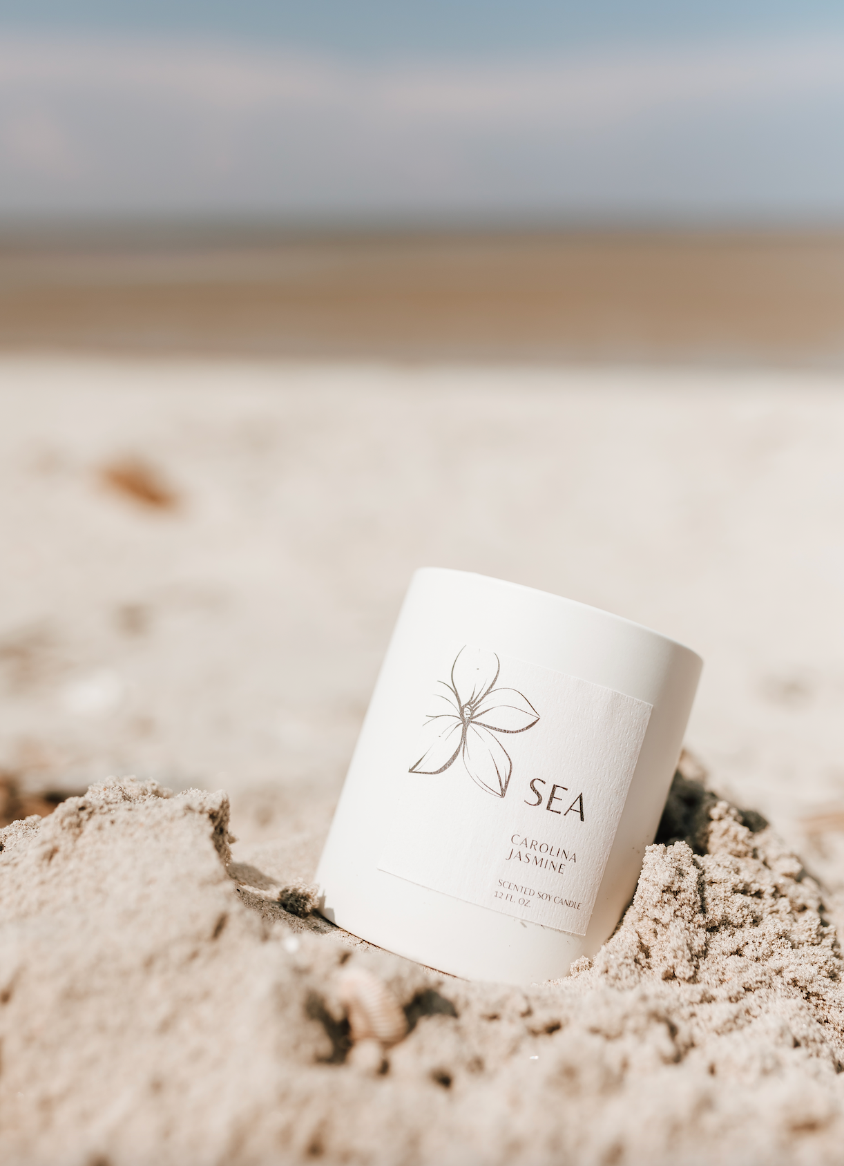 Carolina Jasmine SEA Soy Candle in a ceramic vessel, with notes of rain, sea salt, and jasmine, embedded in sand with a blurred background.