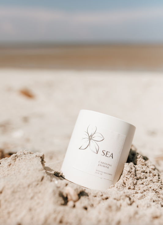 Carolina Jasmine SEA Soy Candle in a ceramic vessel, with notes of rain, sea salt, and jasmine, embedded in sand with a blurred background.