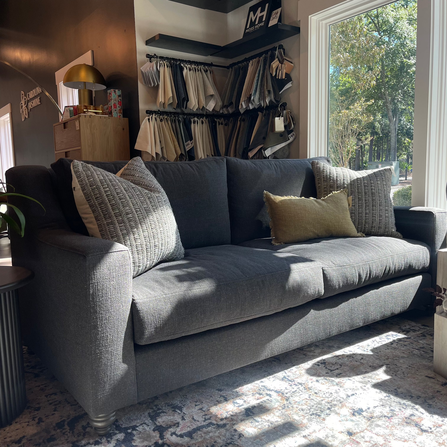 A modern gray Cassie sofa with decorative pillows, placed in a showroom with a variety of fabric samples and lighting on display.